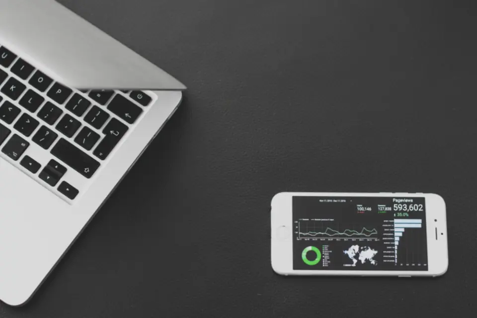 A laptop and a phone on a dark desk, the phone showing a website traffic dashboard with pageview charts, representing behaviour analytics.