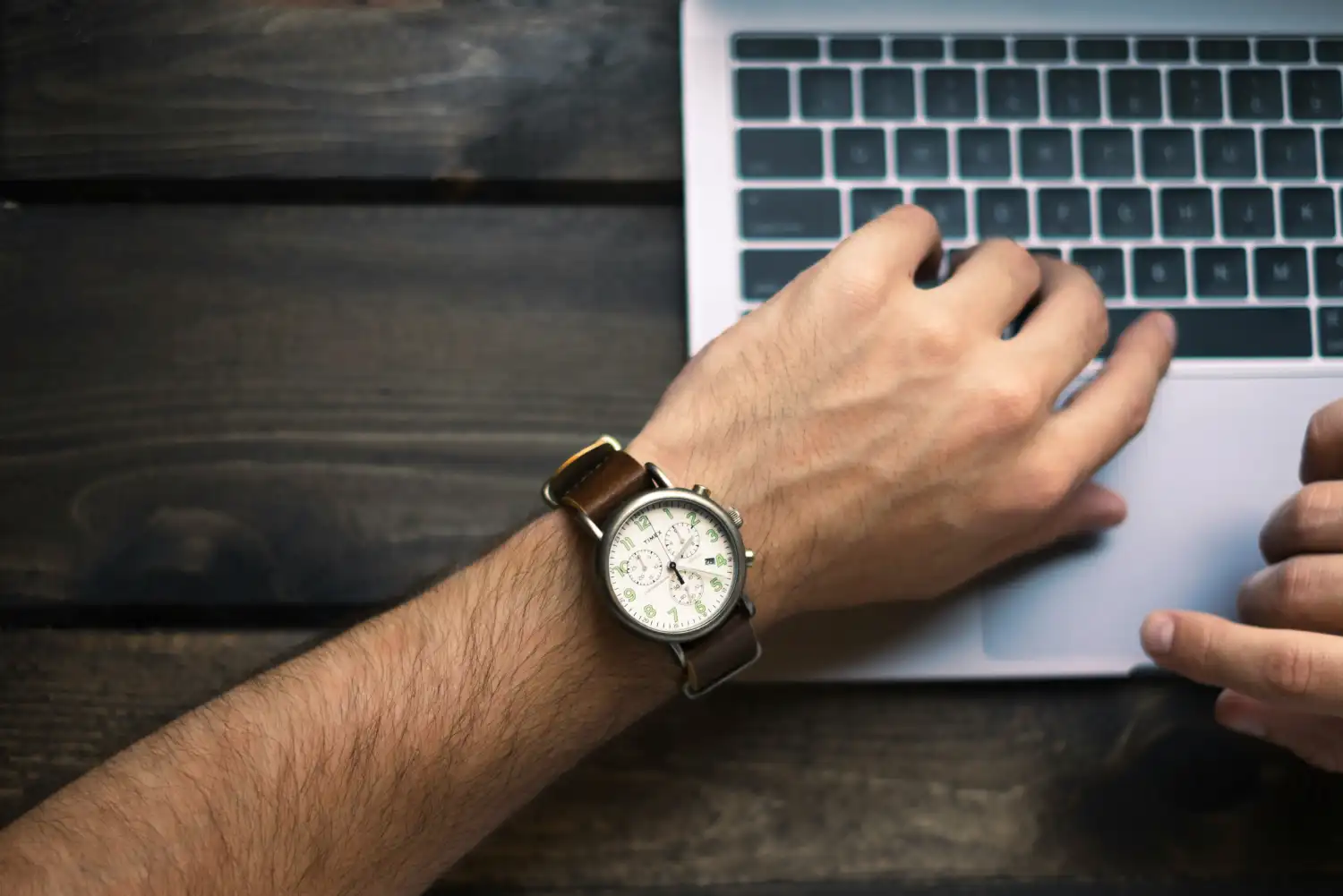 Top-down view of a professional UI/UX designer wearing a classic wristwatch while typing on a laptop, representing the time and craft invested in high-quality human web design.